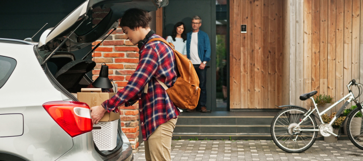 Happy son holding box with his things and positively looking straight at camera. In background mother and dad see their child joining road trip. Young teenager getting ready to drive to college.