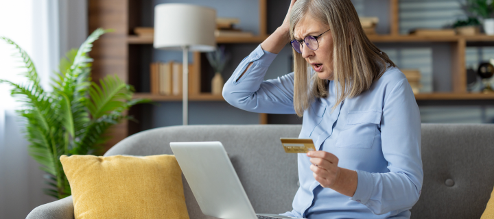 A mature woman looking surprised and anxious while holding a credit card and using a laptop on her sofa at home.